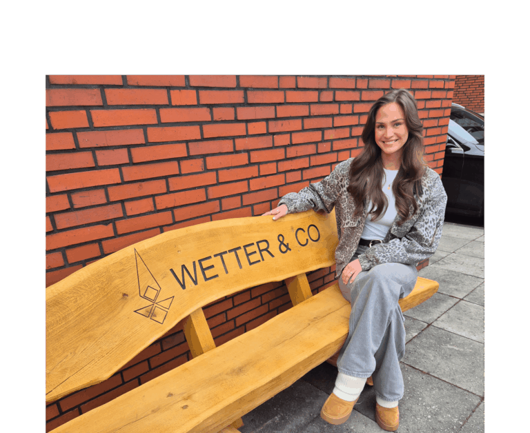 Man sieht Anna Lettow, Mitarbeiterin der Firma Wetter & Co., auf einer Bank sitzen. Anna hat lange braune Haare und lächelt in die Kamera